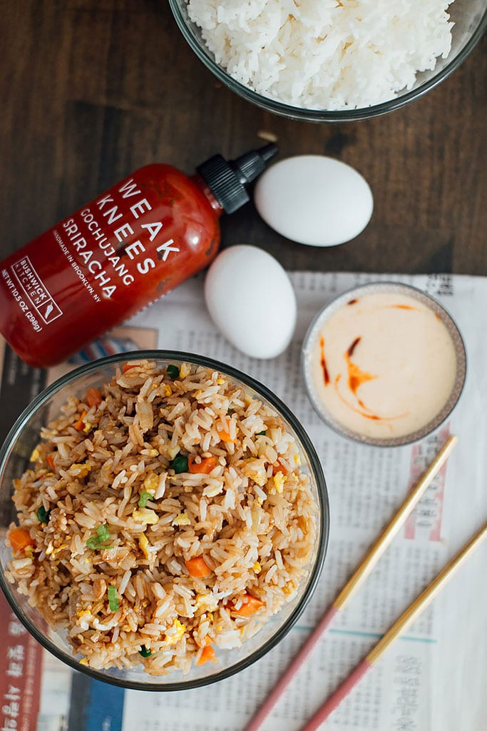 A bowl of hibachi rice on old newspaper with chopsticks, yum yum sauce, srirach sauce, eggs and white rice.