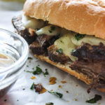 A hearty slow cooker French dip sandwich on white parchment paper with a bowl of au jus dip.