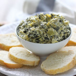 Slow cooker spinach and artichoke dip on a white plate surrounded by sliced French bread.