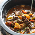 A close up shot of a hearty scoop of slow cooker beef stew in a metal ladle.