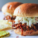 Two slow cooker BBQ pulled chicken burgers topped with coleslaw with pickles and chips.