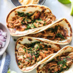 A top down shot of four slow cooker shredded chicken tacos with lime wedges and a dip.