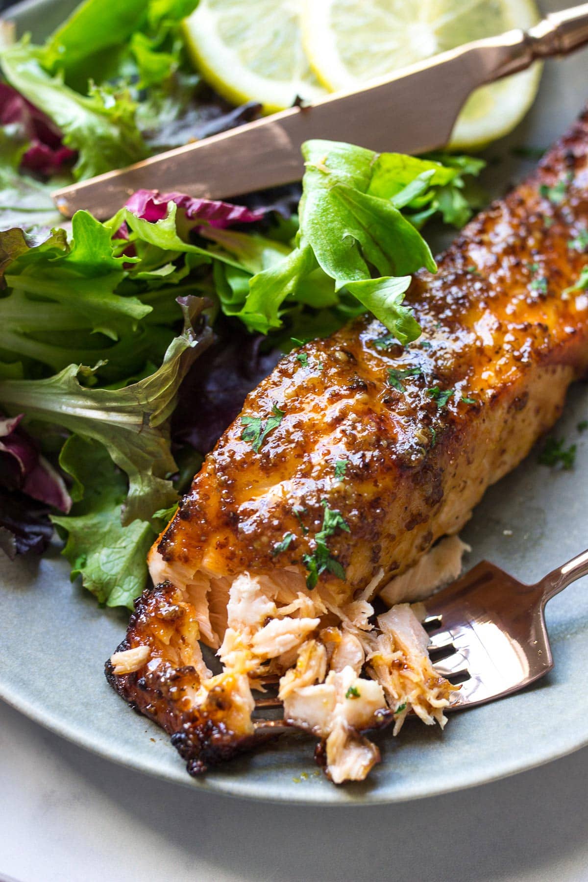 A flaked air fryer honey mustard salmon fillet on a plate with a leafy salad and two lemon slices.