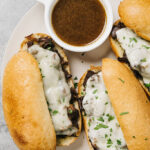 A top down shot of three French dip sandwiches on a white plate with a bowl of au jus.