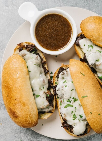 A top down shot of three French dip sandwiches on a white plate with a bowl of au jus.