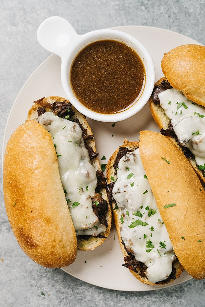 A top down shot of three French dip sandwiches on a white plate with a bowl of au jus.