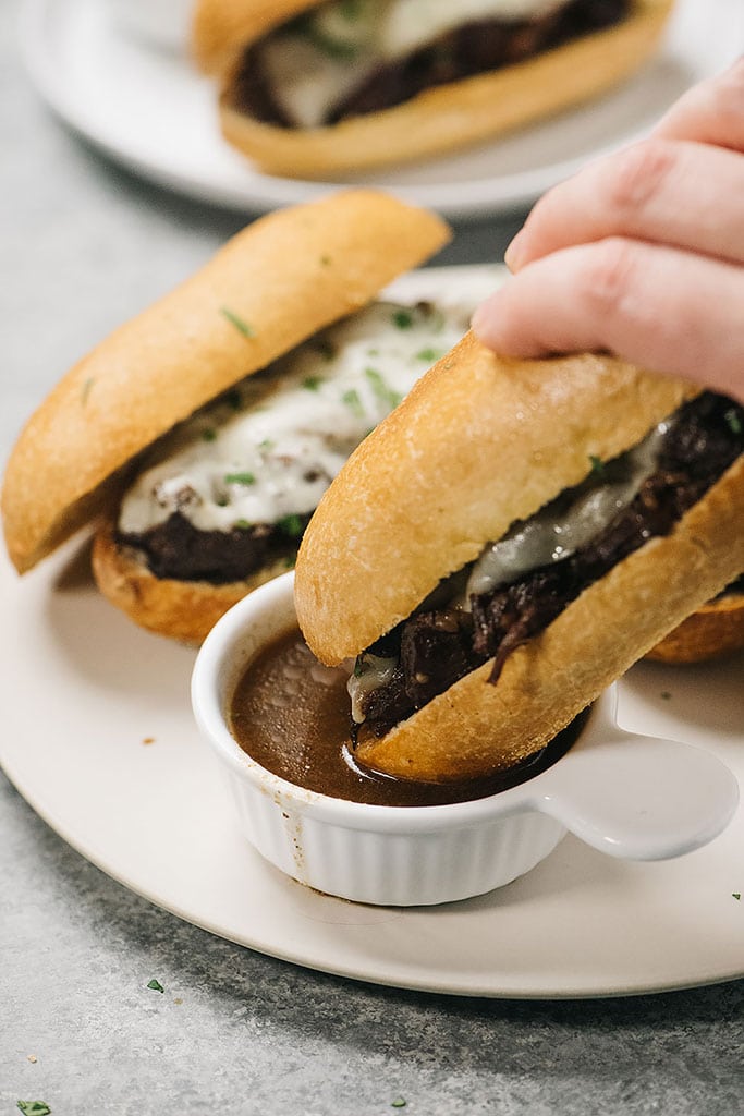 A French dip sandwich being dipped into a bowl of au jus.