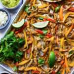 Some chicken fajitas in a sheet pan with onions, bell peppers, cilantro and lime wedges.