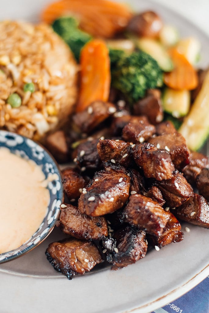 A close up shot of hibachi steak on a plate with hibachi rice, veggies and Yum Yum sauce in the background.