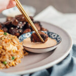 A pair of gold and black chopsticks holding a piece of hibachi steak while dipping into a bowl of Yum Yum sauce.
