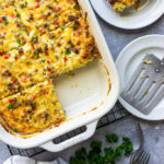 Cheesy sausage hash brown breakfast casserole in a white casserole dish with a slice missing.