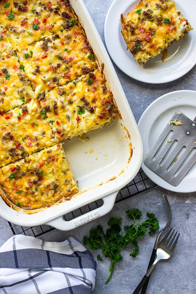 Cheesy sausage hash brown breakfast casserole in a white casserole dish with a slice missing.