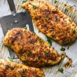 Three freshly baked chicken breasts on parchment paper on a baking sheet.