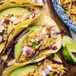 Several chipotle chicken tacos on charred corn tortillas with purple cabbage, avocado, red onions, cilantro and limes.