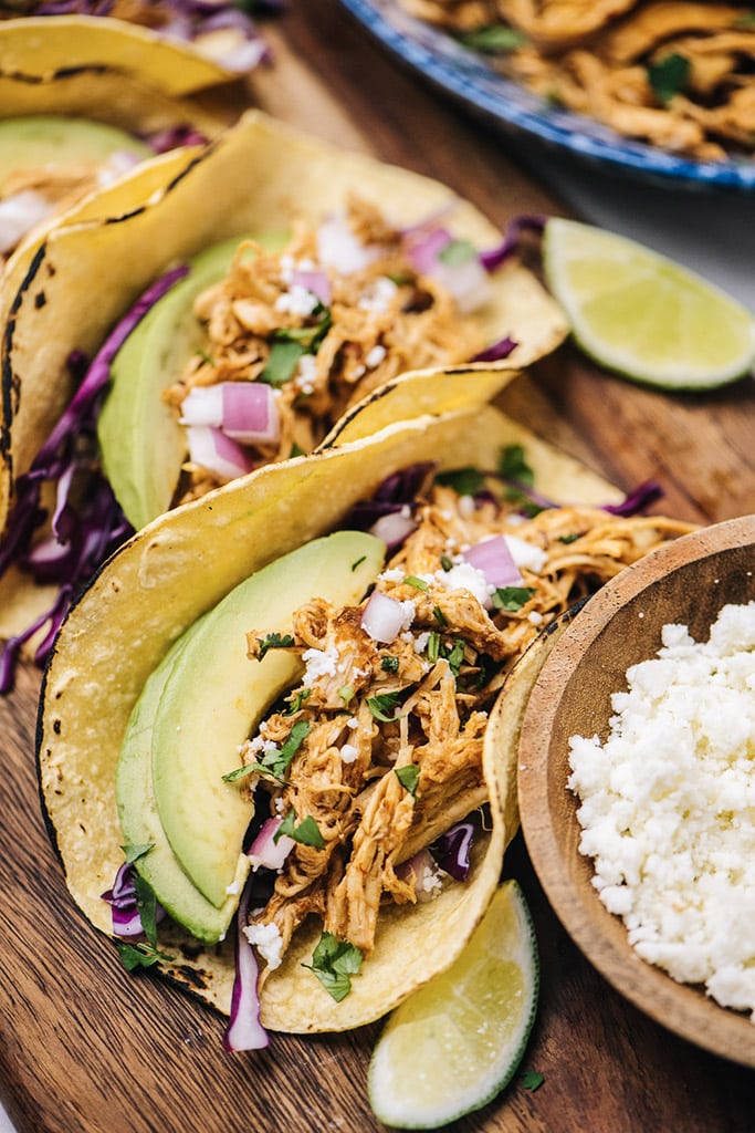 Several chipotle chicken tacos on charred corn tortillas with purple cabbage, avocado, red onions, cilantro, queso fresco and limes.