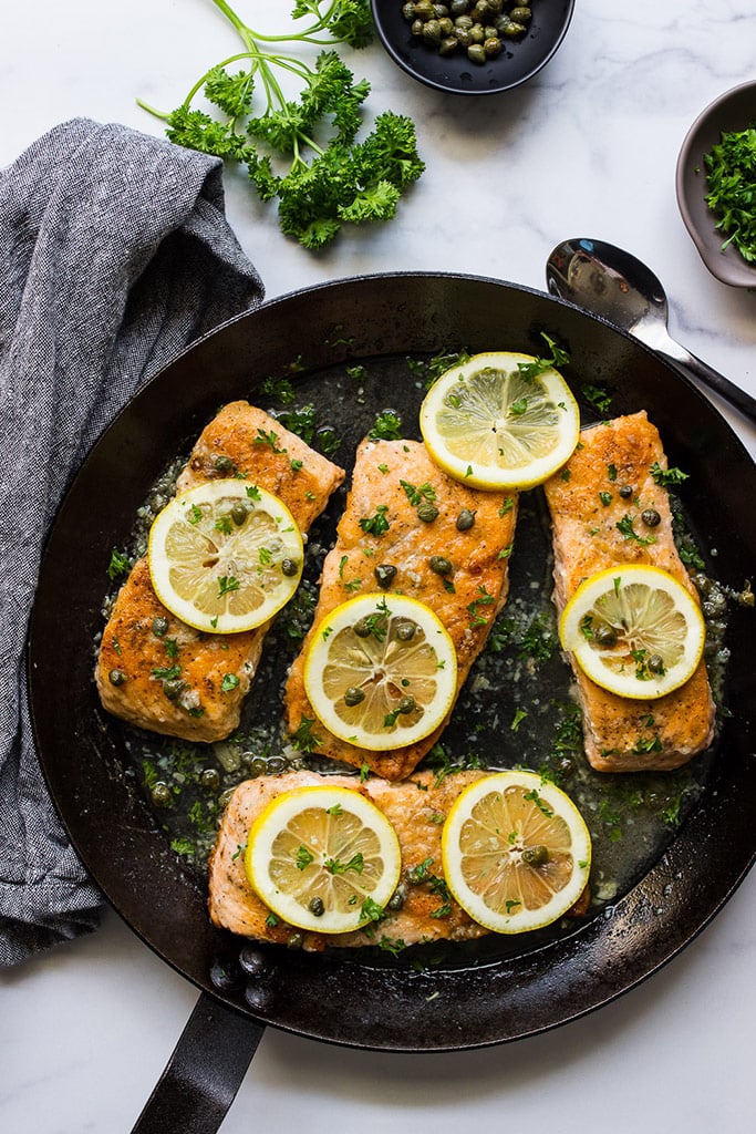 Salmon piccata in a cast iron pan topped with capers, lemon slices and fresh parsley.