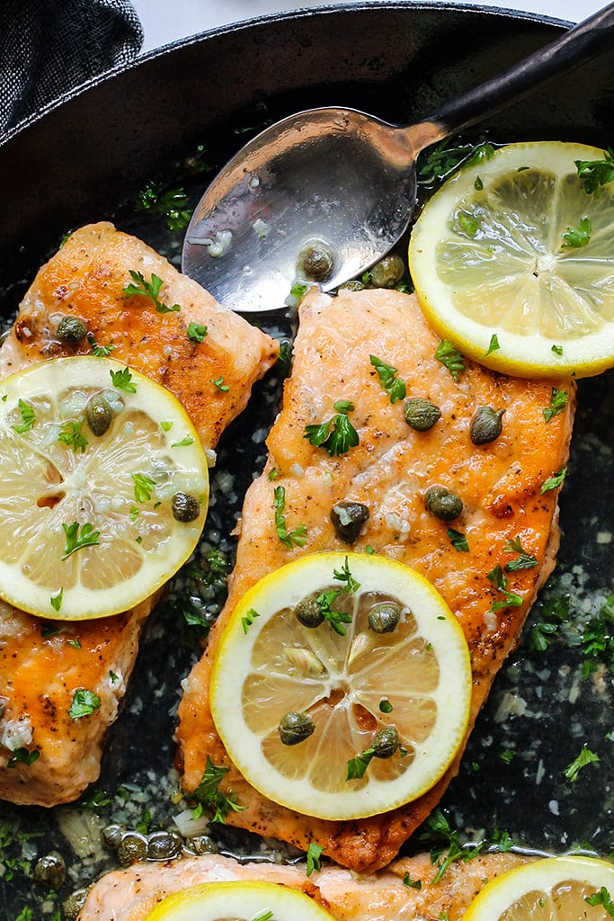 Salmon piccata in a cast iron pan topped with capers, lemon slices and fresh parsley.