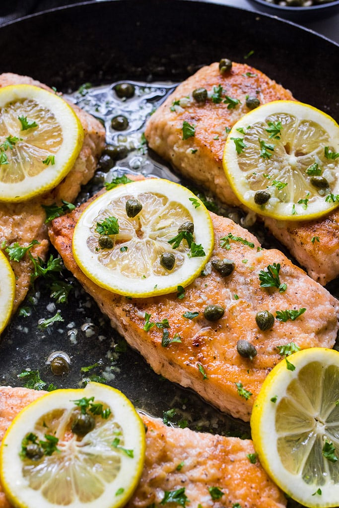 Salmon piccata in a cast iron pan topped with capers, lemon slices and fresh parsley.