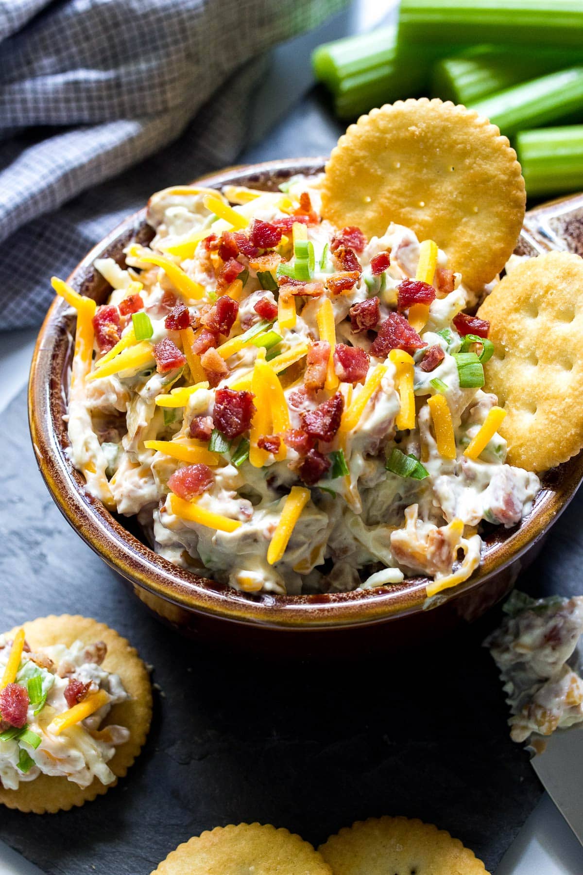 Million dollar dip in a brown bowl with some crackers and topped with shredded cheddar cheese, bacon bits and green onions.