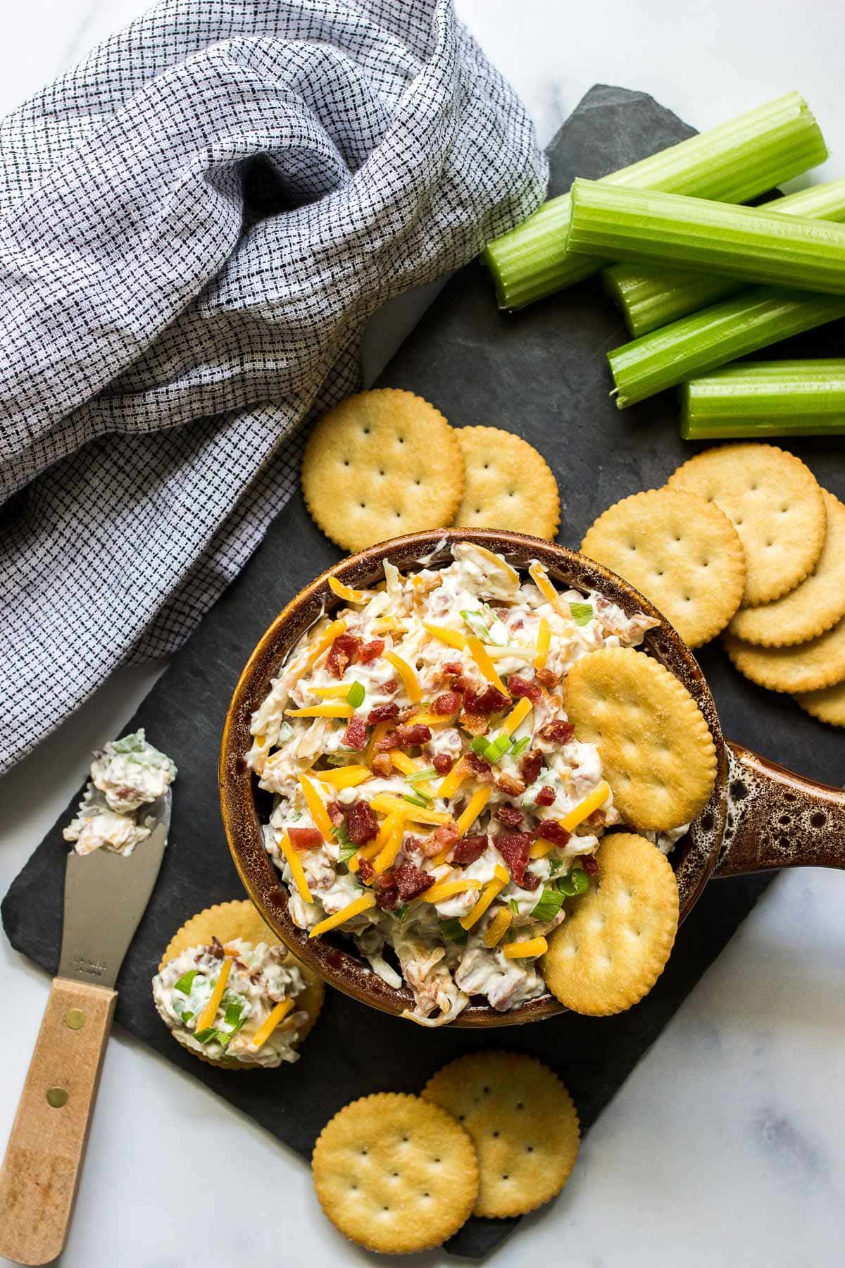 Million dollar dip in a brown bowl with some crackers and topped with shredded cheddar cheese, bacon bits and green onions.