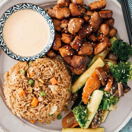 Hibachi rice, chicken and veggies on a white plate with some yum yum sauce on the side.