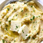 A white bowl of mashed potatoes with slabs of butter and fresh parsley on top.