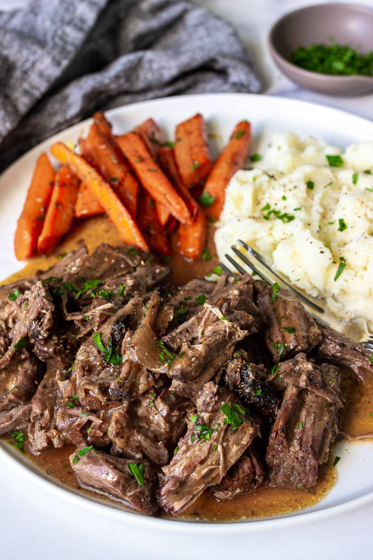 A plate of shredded chuck roast with mashed potatoes and carrots topped with fresh parsley.