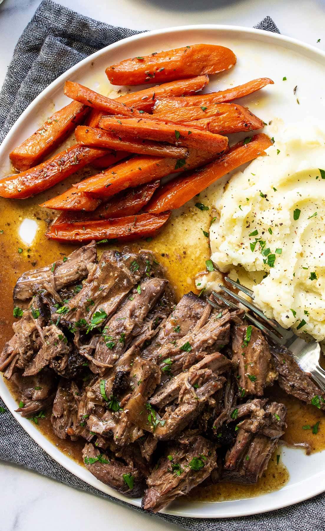 A plate of shredded chuck roast with mashed potatoes and carrots topped with fresh parsley.