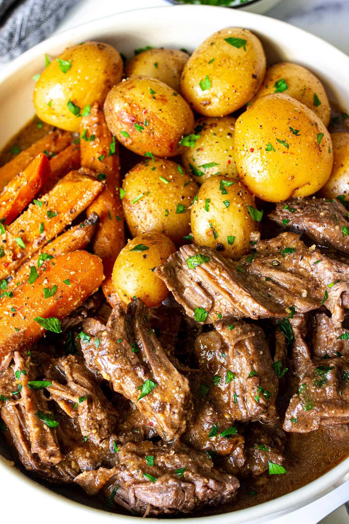 A big bowl of slow cooker pot roast with tender beef, baby yellow potatoes and carrots topped with fresh parsley.