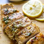 A sliced garlic butter chicken breast on a wooden cutting board with parsley and lemon slices in the background.