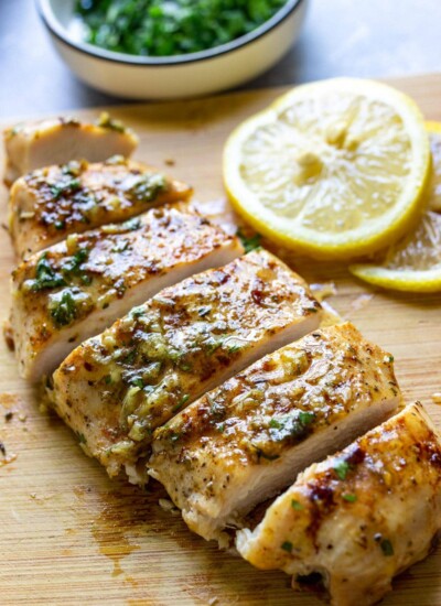 A sliced garlic butter chicken breast on a wooden cutting board with parsley and lemon slices in the background.