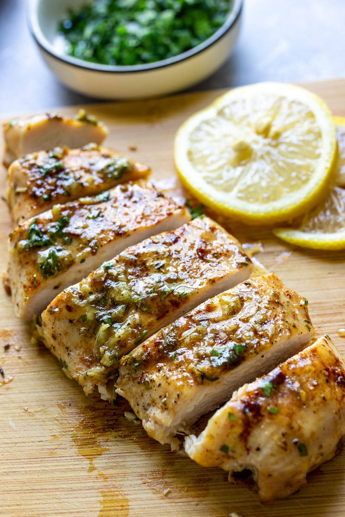 A sliced garlic butter chicken breast on a wooden cutting board with parsley and lemon slices in the background.