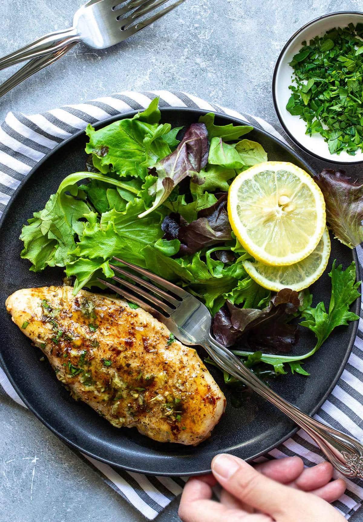 Garlic butter baked chicken breast on a black plate with a mixed salad and lemon slices.