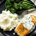 A black plate with creamy garlic butter salmon and broccolini and mashed potatoes on the side.