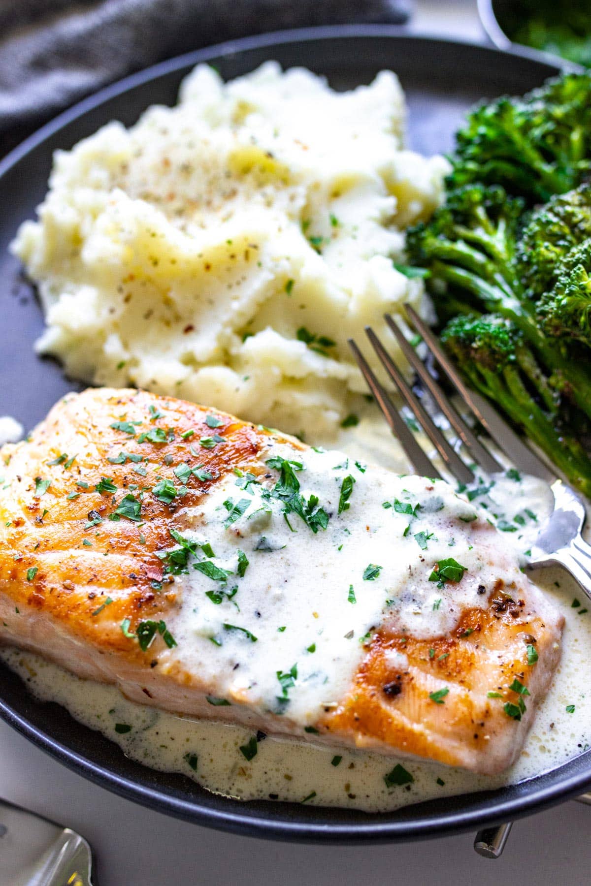 A black plate with creamy garlic butter salmon and broccolini and mashed potatoes on the side.