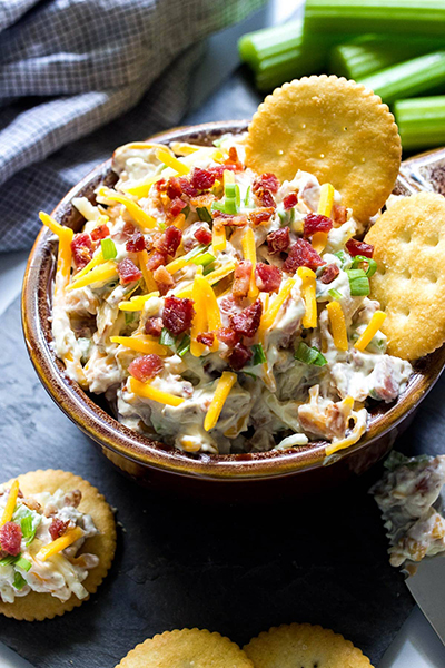 Million dollar dip in a brown bowl with some crackers and topped with shredded cheddar cheese, bacon bits and green onions.