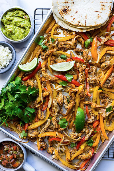 Chicken fajitas on a sheet pan with lime wedges, fresh cilantro, cotija cheese, guacamole and salsa.