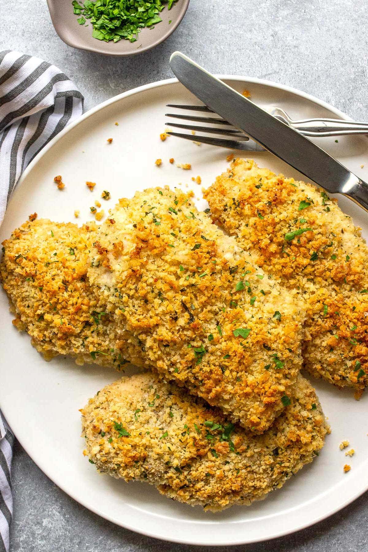 Four baked Parmesan crusted chicken breasts on a white plate topped with parsley.
