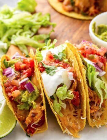 Several cheesy baked chicken tacos stacked together topped with sour cream, tomatos, cilantro, red onions and shredded lettuce.