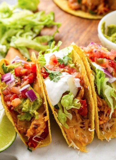 Several cheesy baked chicken tacos stacked together topped with sour cream, tomatos, cilantro, red onions and shredded lettuce.