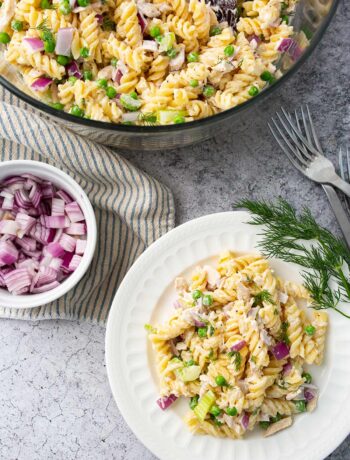 Tuna pasta salad with fresh diced red onions, celery, dill and peas in a mixing bowl and a white plate.