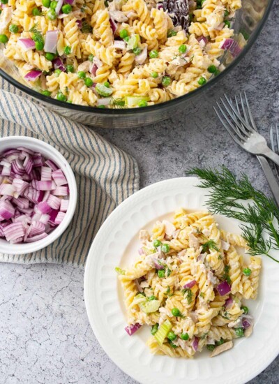Tuna pasta salad with fresh diced red onions, celery, dill and peas in a mixing bowl and a white plate.