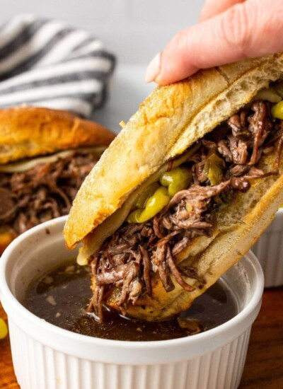 A slow cooker Italian beef sandwich being dipped into a bowl of au jus.