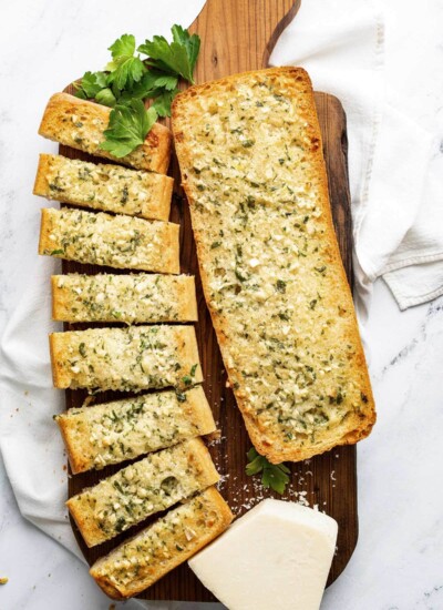 Homemade garlic bread on a cutting board topped with freshly chopped parsley and grated cheese.