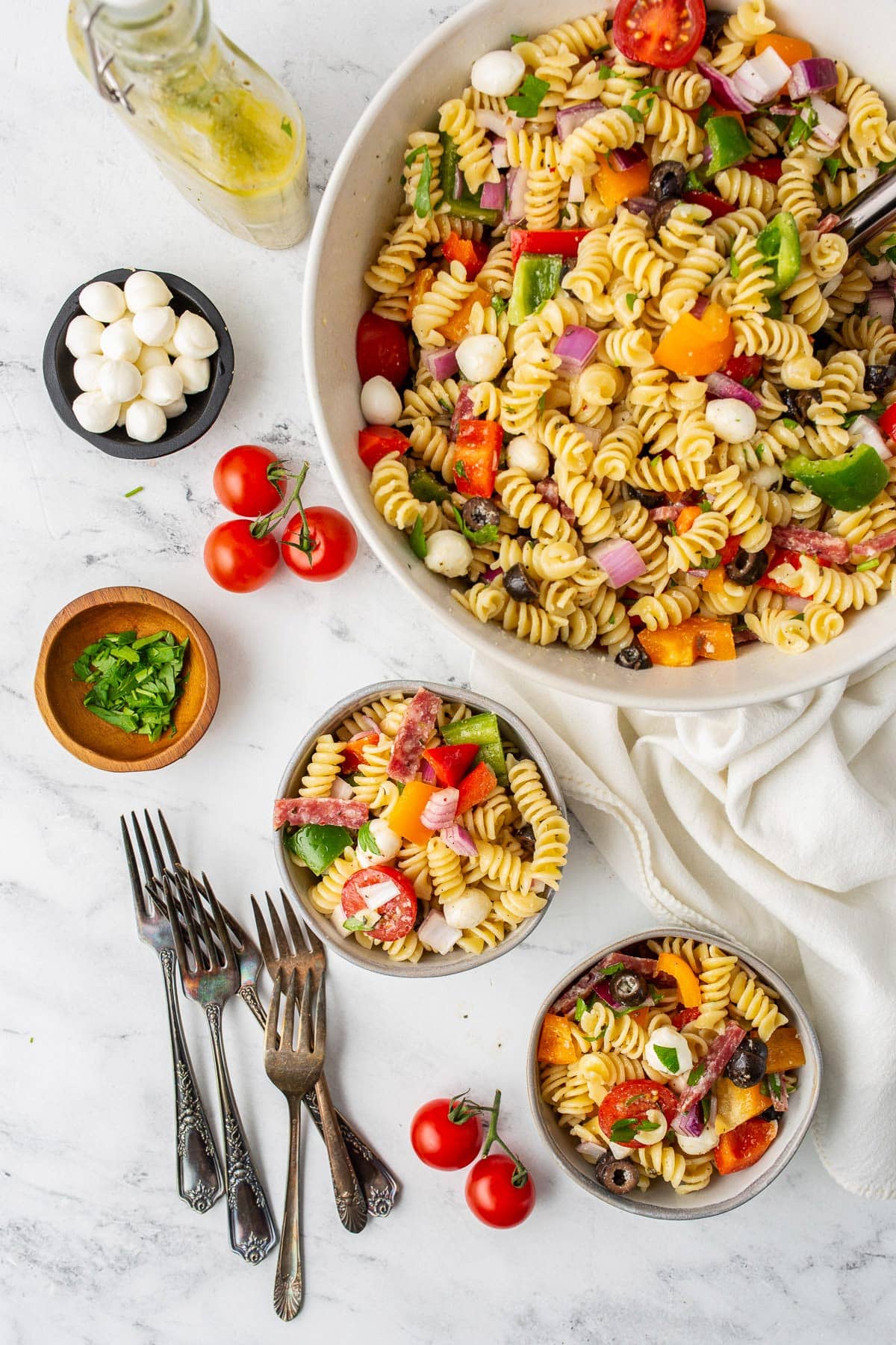 Italian pasta salad in several white bowls surrounded by tomatoes on the vine, chopped parsley, mozzarella balls and dressing.