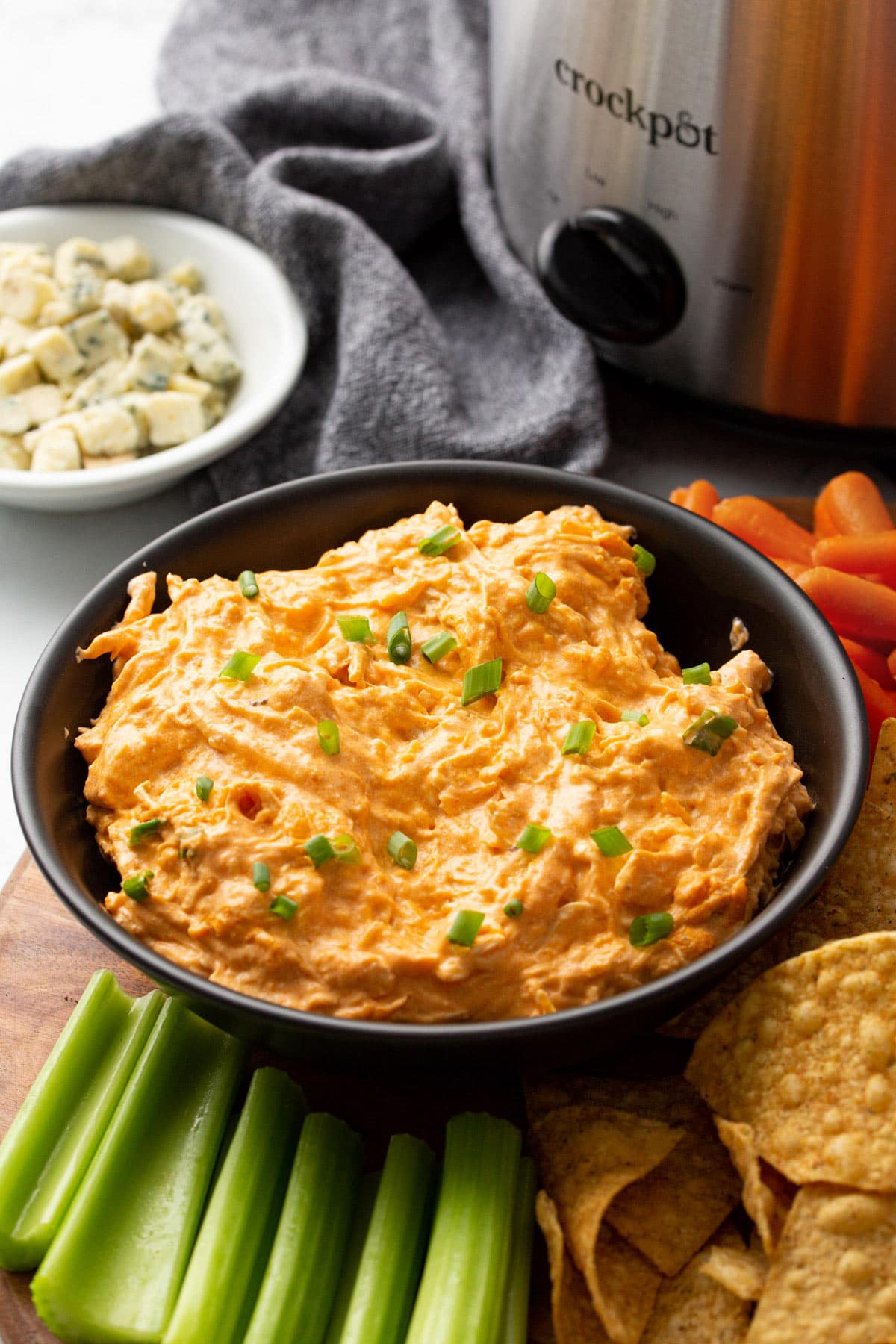 Slow-cooked buffalo chicken served in a clay pot and a black bowl surrounded by tortilla chips, crackers, celery sticks, baby carrots and blue cheese crumbs.