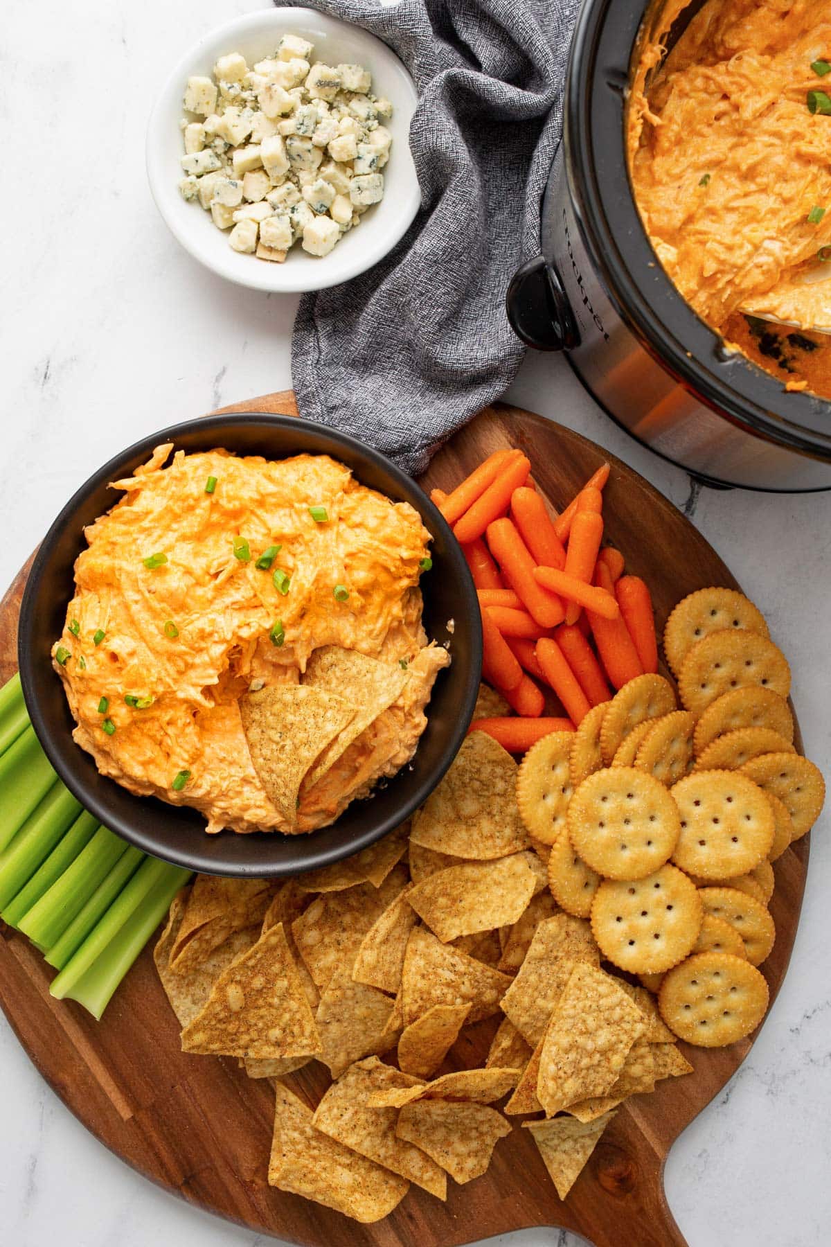 Slow cooker buffalo chicken served in a crock pot and black bowl surrounded by tortilla chips, crackers, celery sticks, baby carrots and blue cheese crumbles.