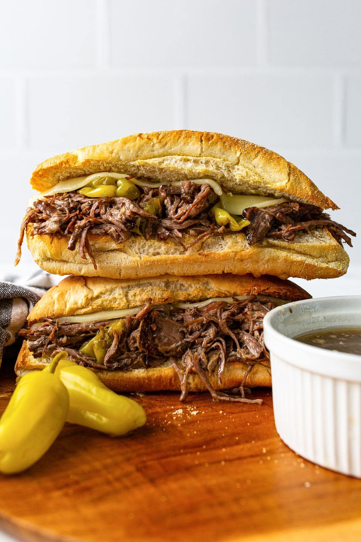 Two slow cooker Italian beef sandwiches stacked on each other with pepperoncini peppers and a bowl of au jus.