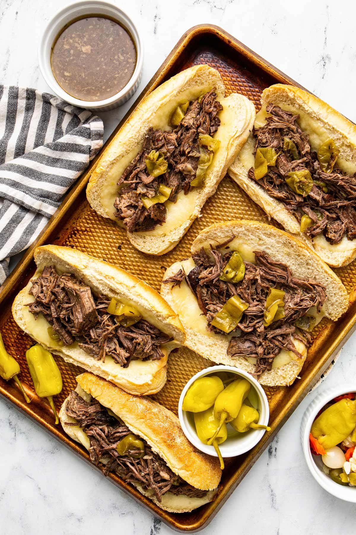 Several slow cooker Italian beef sandwiches on a baking sheet with pepperoncini peppers and a bowl of au jus.