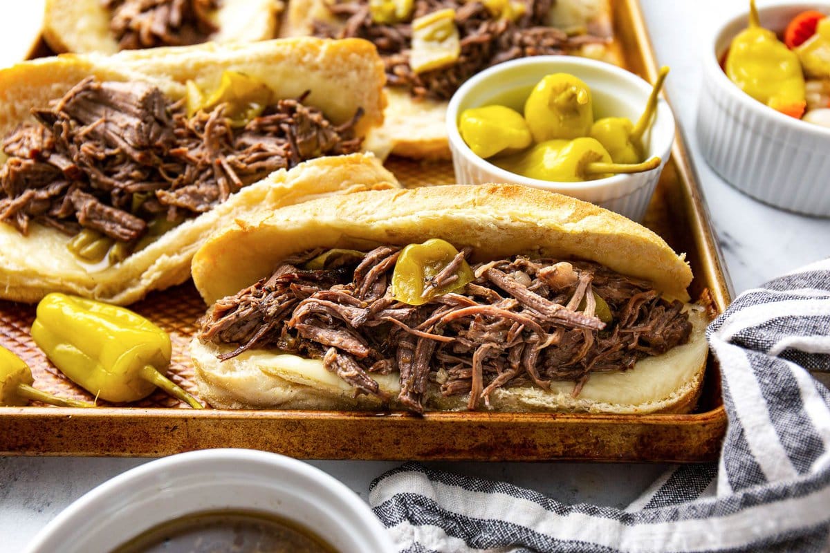 Several slow cooker Italian beef sandwiches on a baking sheet with pepperoncini peppers and a bowl of au jus.